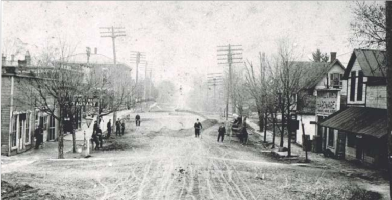 Old view of Ostrander, a dirt road lined with old buildings and power lines, people and horse carriages stand on the sides of the street