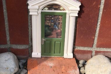 A mini green door placed against a brick wall with a little window peering into a make believe room full of books