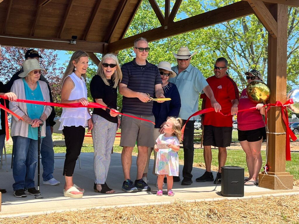 A group of happy people cutting a large, red ribbon with big, gold scissors.