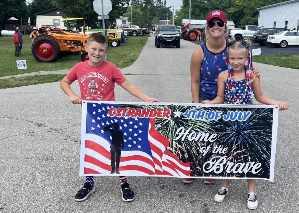 Two kids and an adult woman dressed in 4th of July attire posing for a photo with the 4th of July event banner