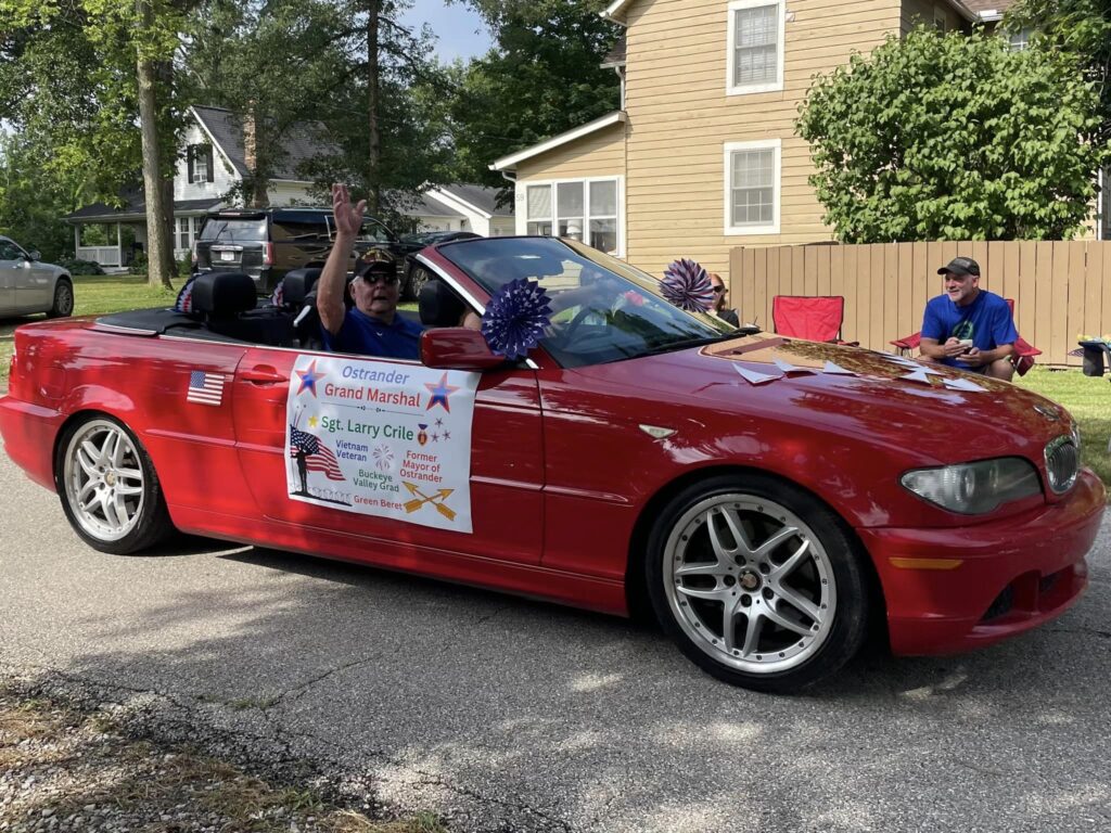 Ostrander Grand Marshal, Larry Crile, smiles and waves in his red BMW decorated with 4th of July decore