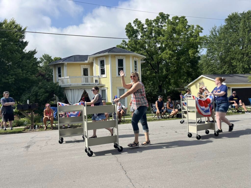 Smiling and waving women pushing book carts in the 4th of July parade