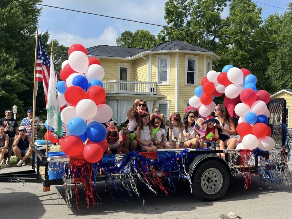 Kids and adults wearing red, white and blue attire riding on a float decorated with patriotic balloons